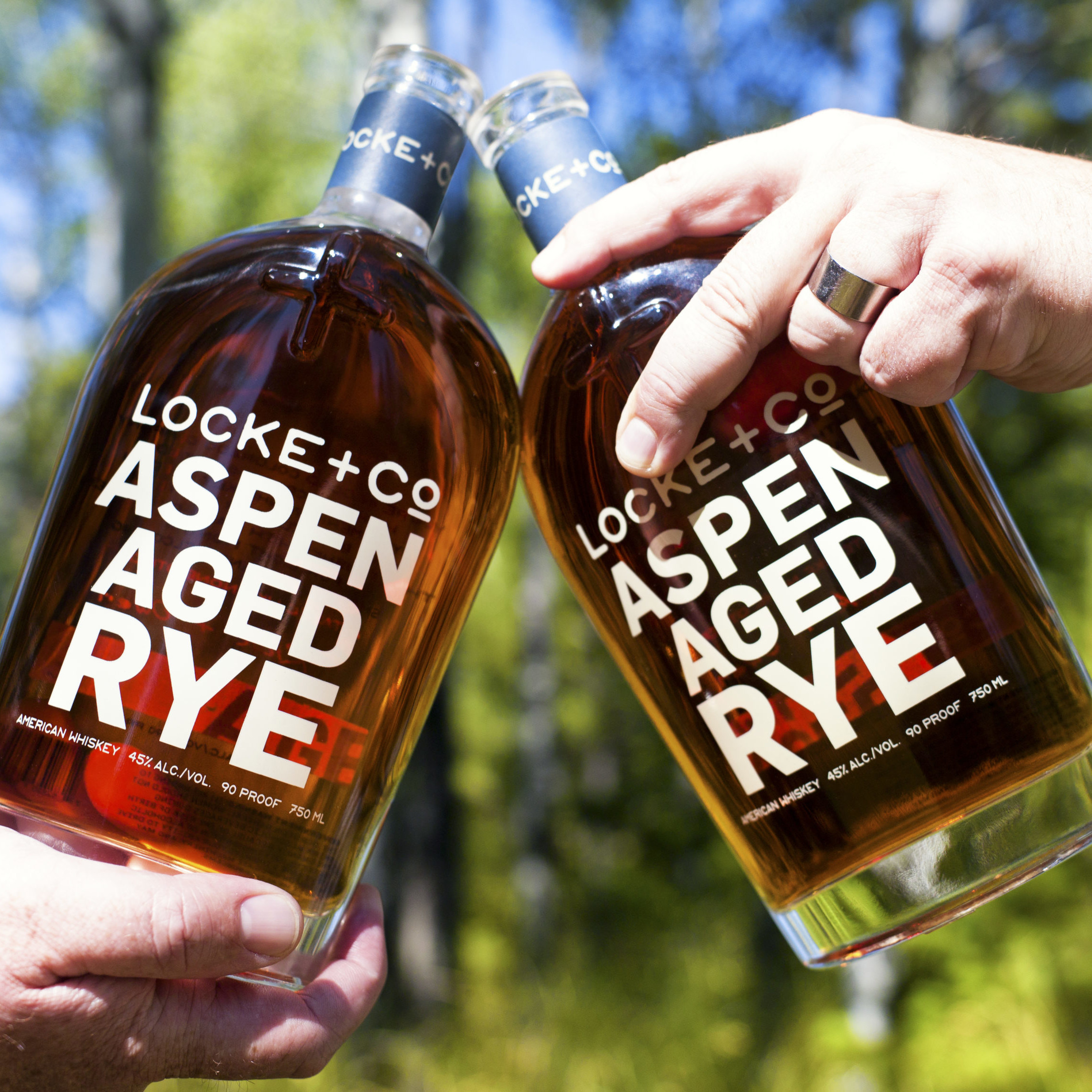 Two people cheer-sings Locke + Co Distilling Aspen Aged Rye Whiskey Bottles