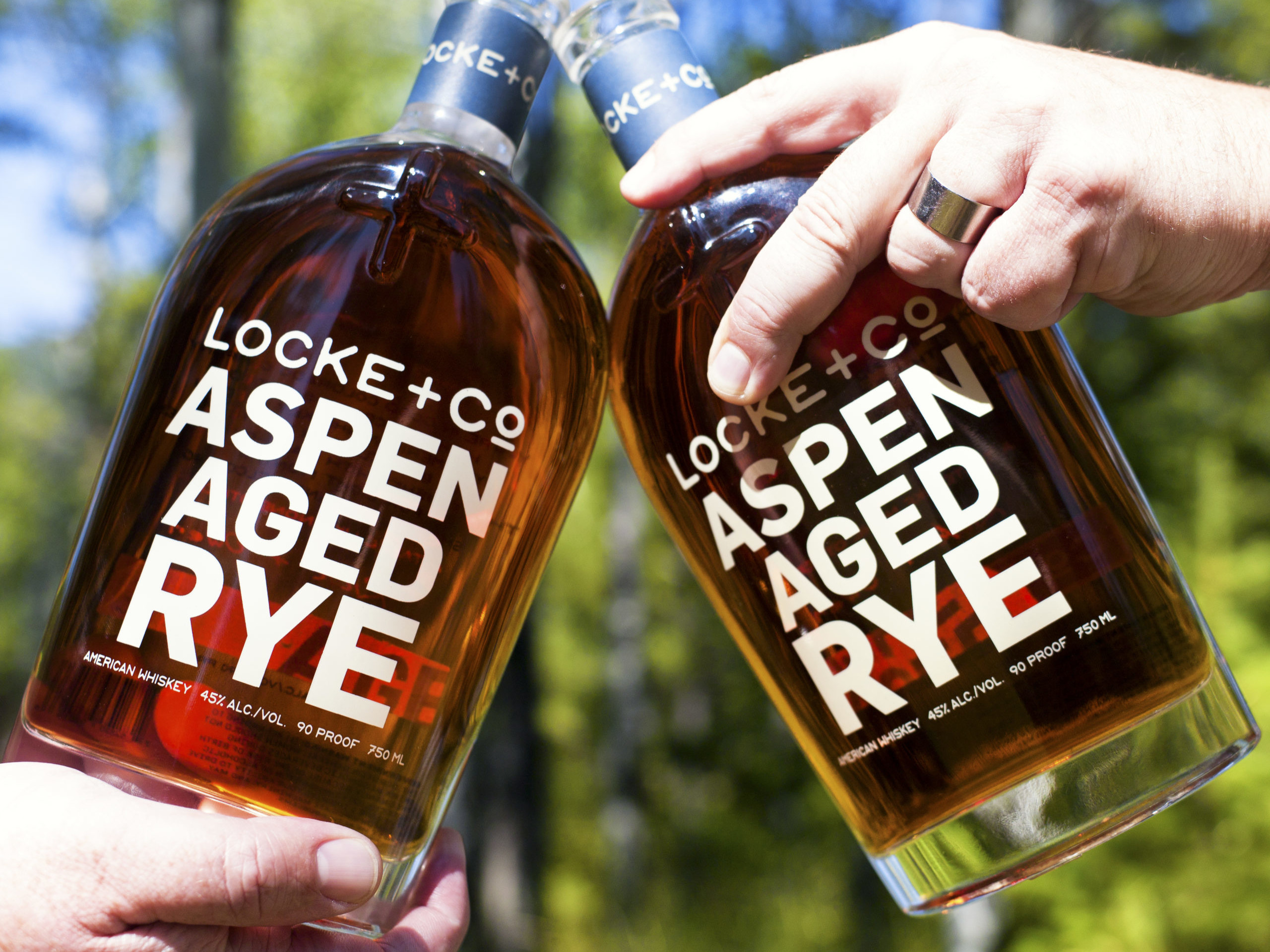Two people cheer-sings Locke + Co Distilling Aspen Aged Rye Whiskey Bottles