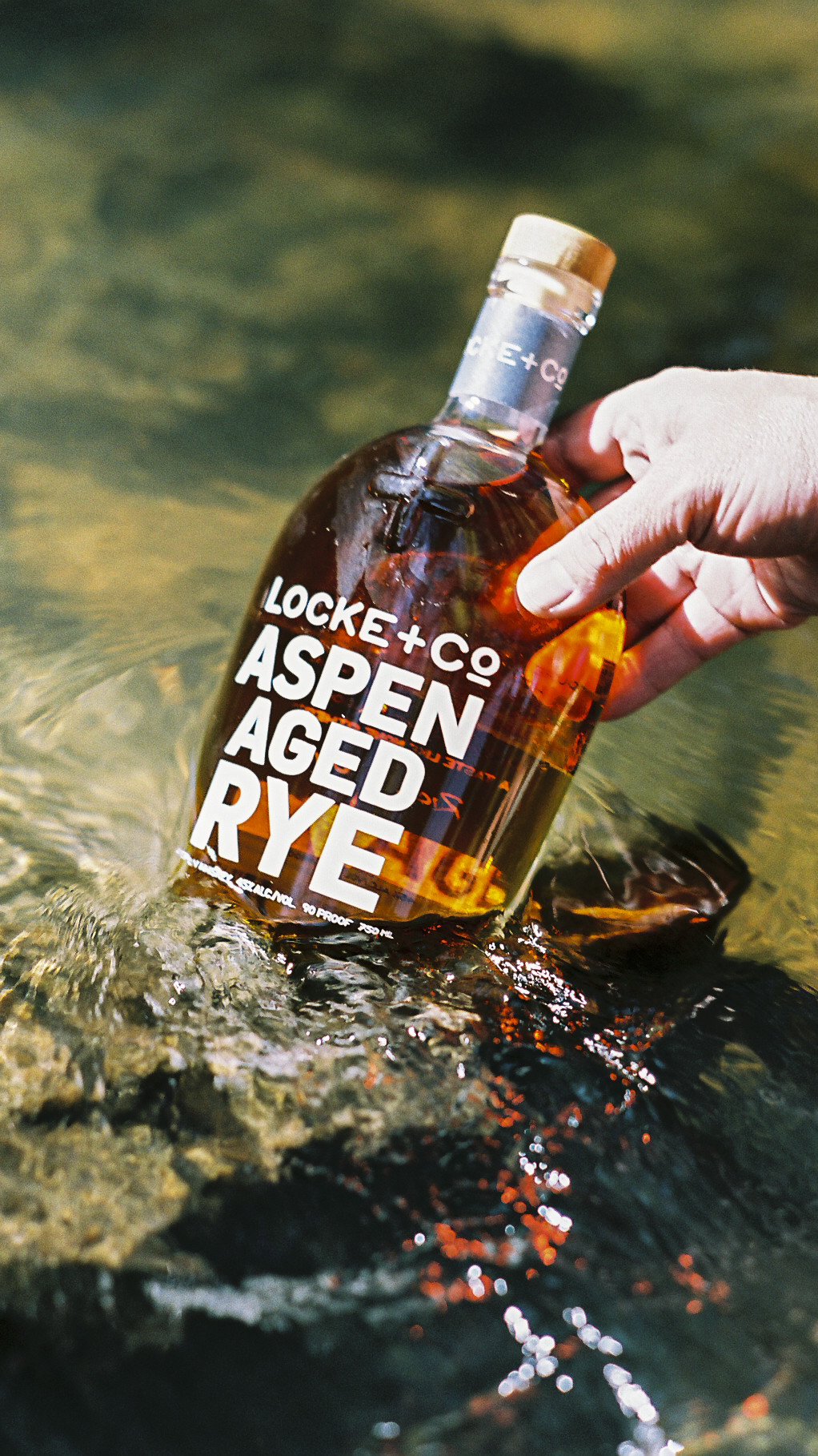 A hand holding a Locke + Co. Aspen Aged Rye Whiskey Bottle in a trout river