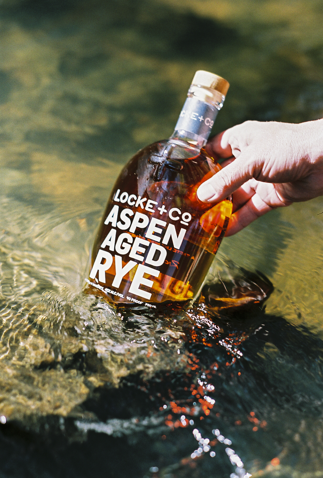 A hand holding a Locke + Co. Aspen Aged Rye Whiskey Bottle in a trout river