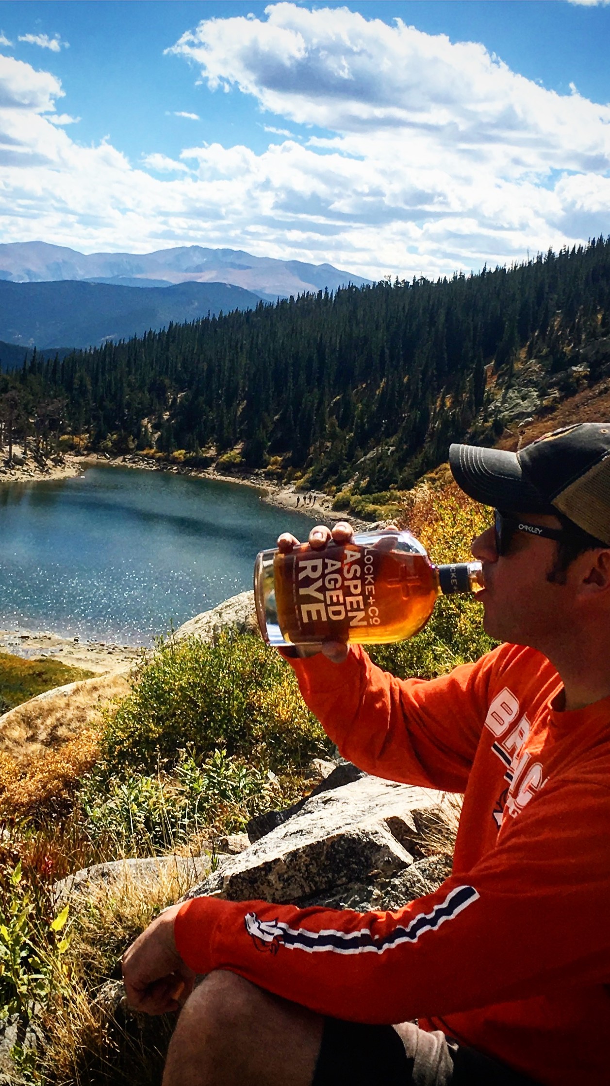 Rick Talley drinking out of the Locke + Co. Aspen Aged Rye Whiskey Bottle next to a mountain lake