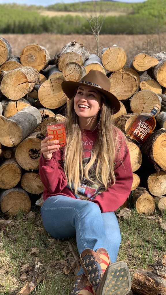 Happy women holding a whiskey cocktail sitting on the ground in front of a stack of aspen logs with a Locke + Co. Aspen Aged Rye Whiskey Bottle