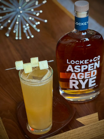 Celebration Rye Cocktail with apple cubes on a toothpick garnishes sitting on a wood coaster in front of a bottle of Locke + Co. Aspen Aged Rye Whiskey on a wood table