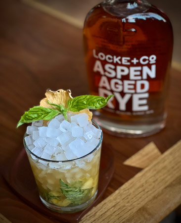 Pineapple Basil Smash with basil and dried pineapple garnishes sitting on a wood coaster in front of a bottle of Locke + Co. Aspen Aged Rye Whiskey on a wood table