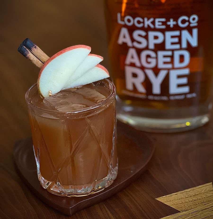 Apple Cider Old Fashioned with apple and cinnamon stick garnishes sitting on a wood coaster in front of a bottle of Locke + Co. Aspen Aged Rye Whiskey on a wood table