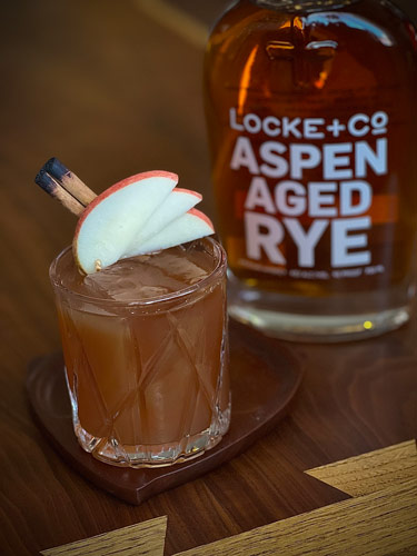 Apple Cider Old Fashioned with apple and cinnamon stick garnishes sitting on a wood coaster in front of a bottle of Locke + Co. Aspen Aged Rye Whiskey on a wood table