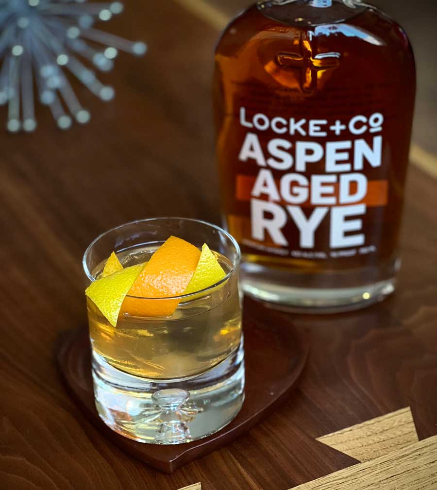 Old Fashioned sitting on a wood coaster in front of a bottle of Locke + Co. Aspen Aged Rye Whiskey on a wood table with a silver mid century sputnik in the background
