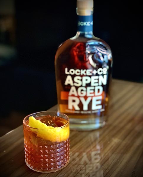 Needle In The Hay Cocktail and a bottle of Locke + Co. Aspen Aged Rye Whiskey on a wood table