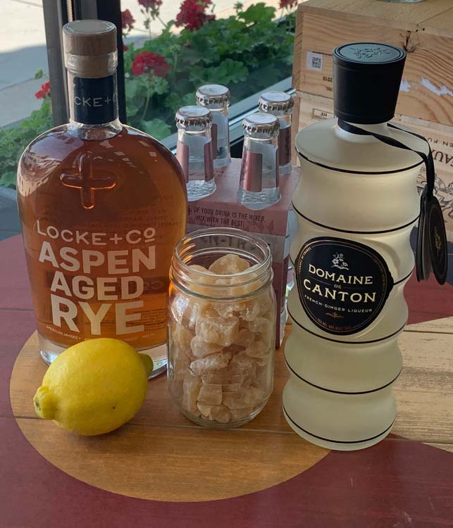 Bottle of Locke + Co. Aspen Aged Rye Whiskey, Bottle of Domaine de Canton Ginger Liqueur, Mason Jar of Candied Ginger & Lemon in from of a 4 pack of Fever Tree Soda Water on a wood table with the image of the colorado flag