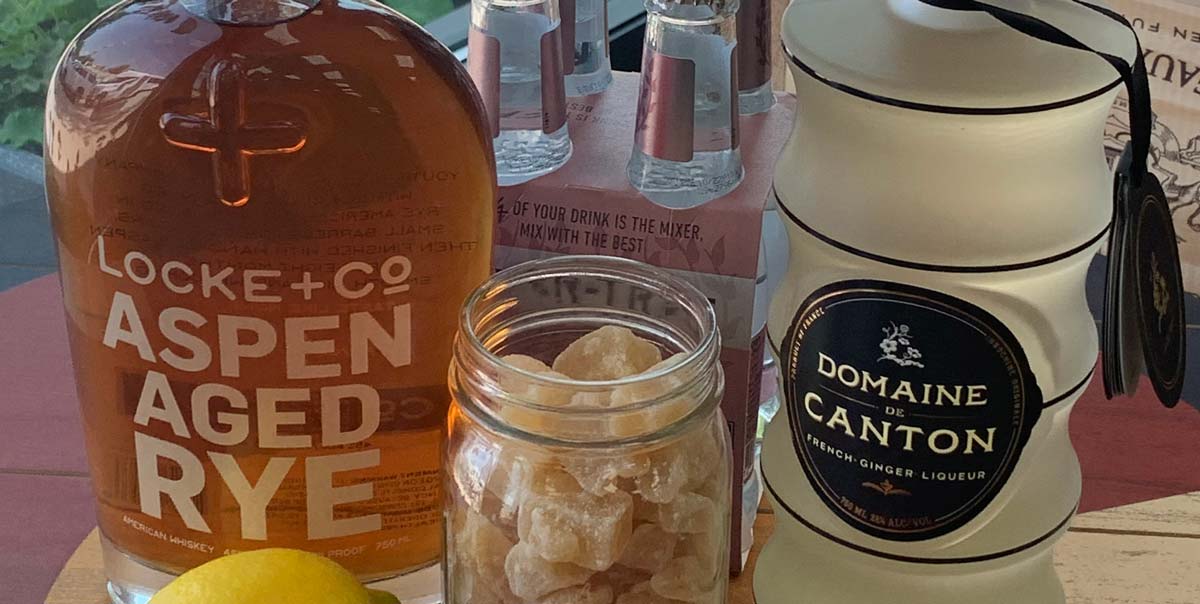 Bottle of Locke + Co. Aspen Aged Rye Whiskey, Bottle of Domaine de Canton Ginger Liqueur, Mason Jar of Candied Ginger & Lemon in from of a 4 pack of Fever Tree Soda Water on a wood table with the image of the Colorado flag