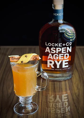 apple cider hot toddy and a bottle of Locke & Co. Aspen Aged Rye Whiskey on a wood table with a black background