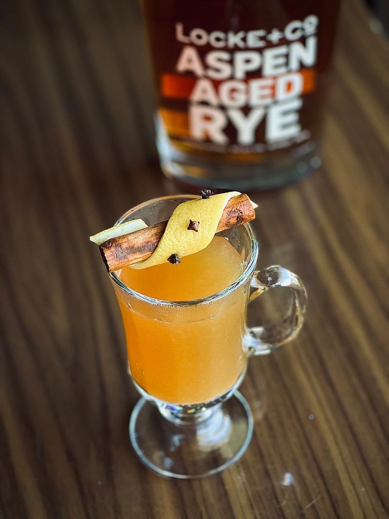 apple cider hot toddy and a bottle of Locke & Co. Aspen Aged Rye Whiskey on a wood table