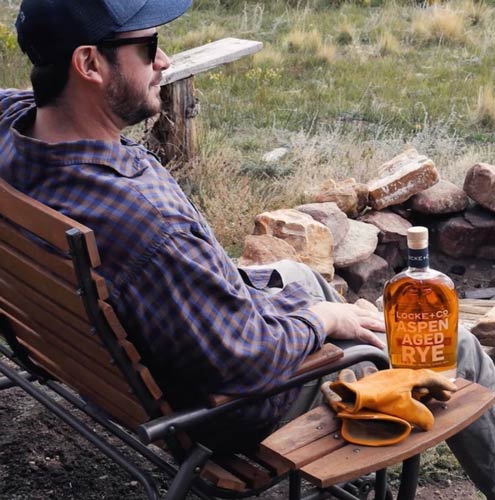 Co-Founder Rick Talley sitting outside in chair in front of a fire-pit with a bottle of Locke + Co Aspen Aged Rye Whiskey
