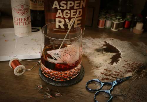 RepYourWater Black Manhattan Cocktail on a table with lure making materials, Locke + Co Aspen Aged Rye Whiskey & a bottle of Fee Brothers Cherry Bitters