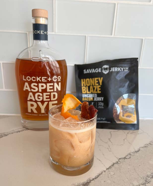 Bacon, Egg & Whiskey Cocktail on counter with a bottle of Locke + Co. Aspen Aged Ryes Whiskey & a Bag of Savage Honey Blaze Bacon