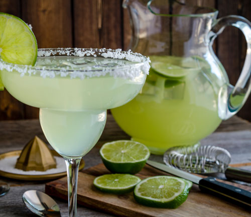 Table filled with glasses and pitcher filled with classic lime margarita cocktails, fresh limes and barware