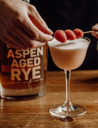 locke+club cocktail with three raspberries on pick laid over top of glass and a bottle of Locke + Co Aspen Aged Rye Whiskey