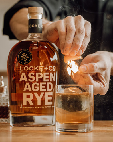 the centennial cocktail with an orange flame twist and a bottle of Locke + Co Aspen Aged Rye Whiskey