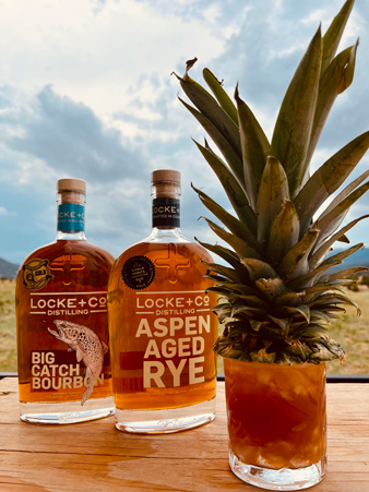 Two bottles of Locke + Co whiskey—Big Catch Bourbon and Aspen Aged Rye—sitting on a wooden table next to a tropical-style cocktail topped with a whole pineapple, with a scenic outdoor view and cloudy sky in the background.