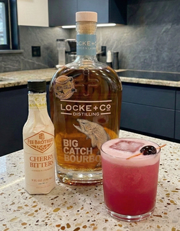 Cherry Whiskey Sour cocktail with egg white foam and cherry garnish served in a rocks glass, pictured with Big Catch Bourbon and cherry bitters on a kitchen counter.